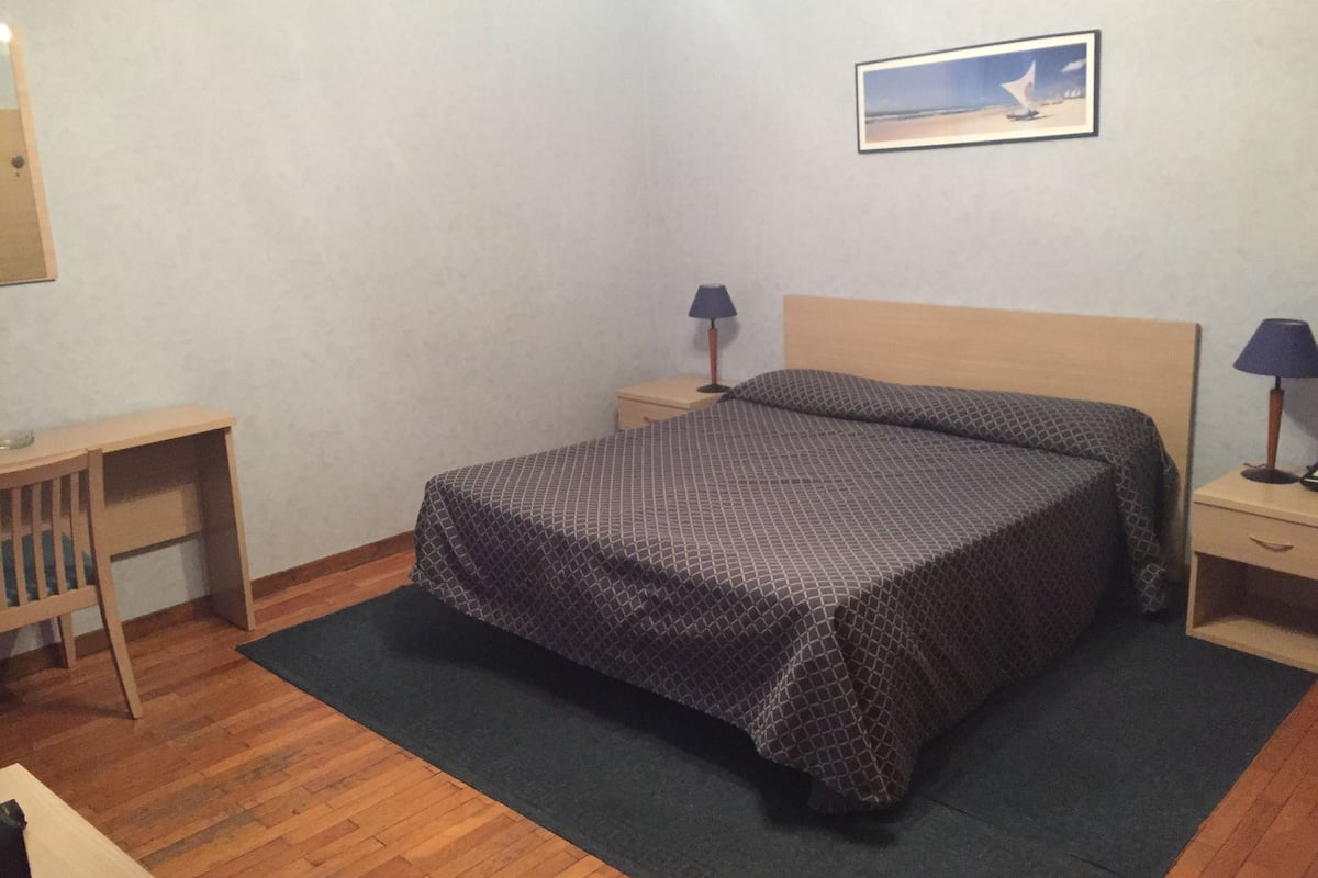 A bedroom with a bed, two bedside tables, a nightstand, a wooden dresser, a dark rug, and a framed picture on the wall.
