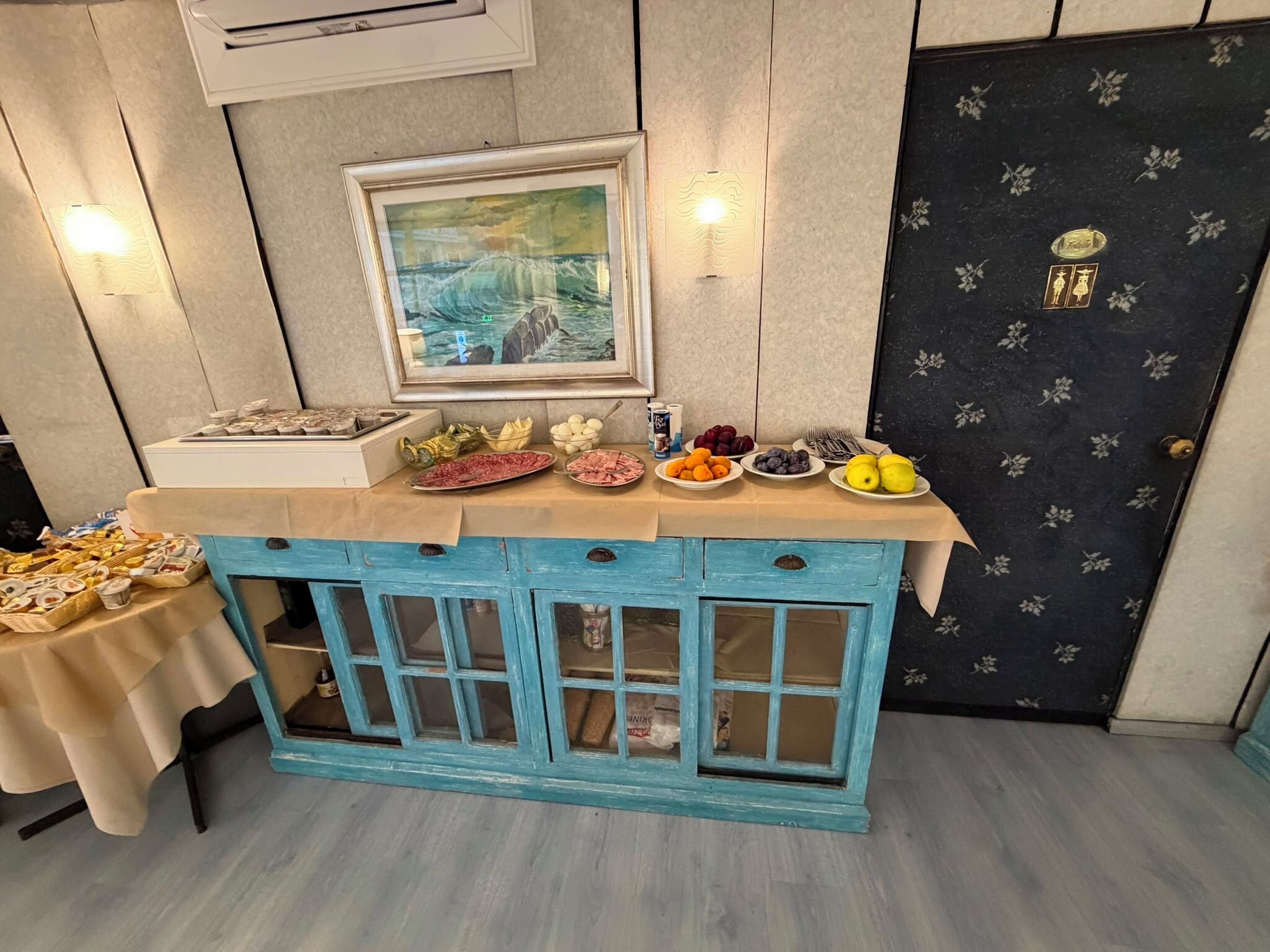 Een kamer met een smaragdgroene kast met glazen deuren, een lijstwerk schilderij aan de muur en verschillende etenswaren op het aanrecht.
