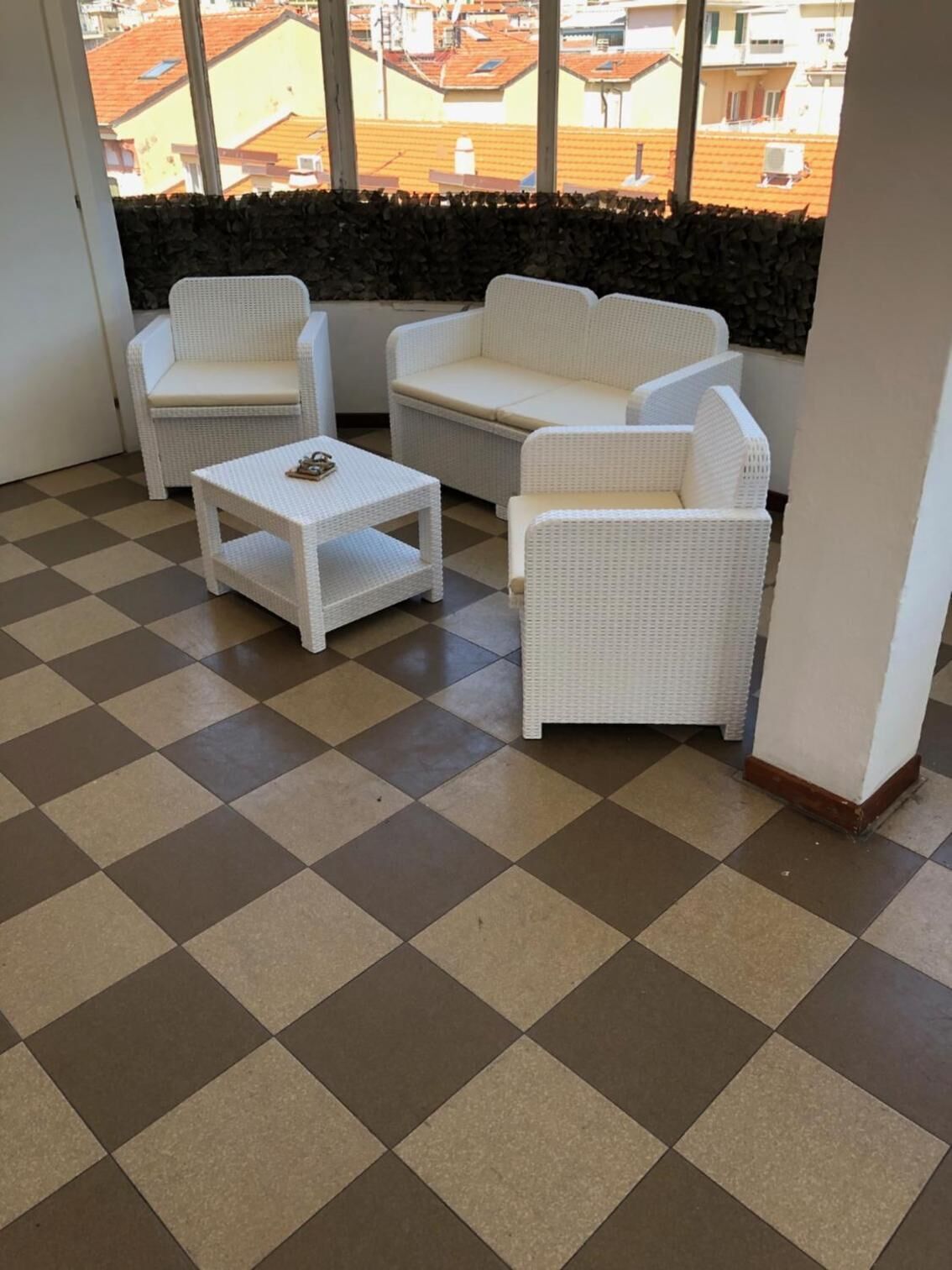 A balcony with a checkered floor, white wicker furniture, and a view of buildings through a window.