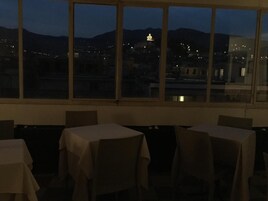 Un comedor con mesas y sillas, ventanas amplias con vistas a un paisaje urbano y montañas al anochecer.