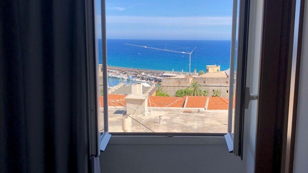 Ein Fenster mit Blick auf eine Küstenlandschaft, darunter ein Yachthafen und Gebäude mit orangefarbenen Dächern.