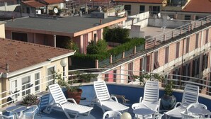 Ein Poolbereich auf dem Dach mit Liegestühlen und Tischen, umgeben von Gebäuden mit roten Ziegeldächern.