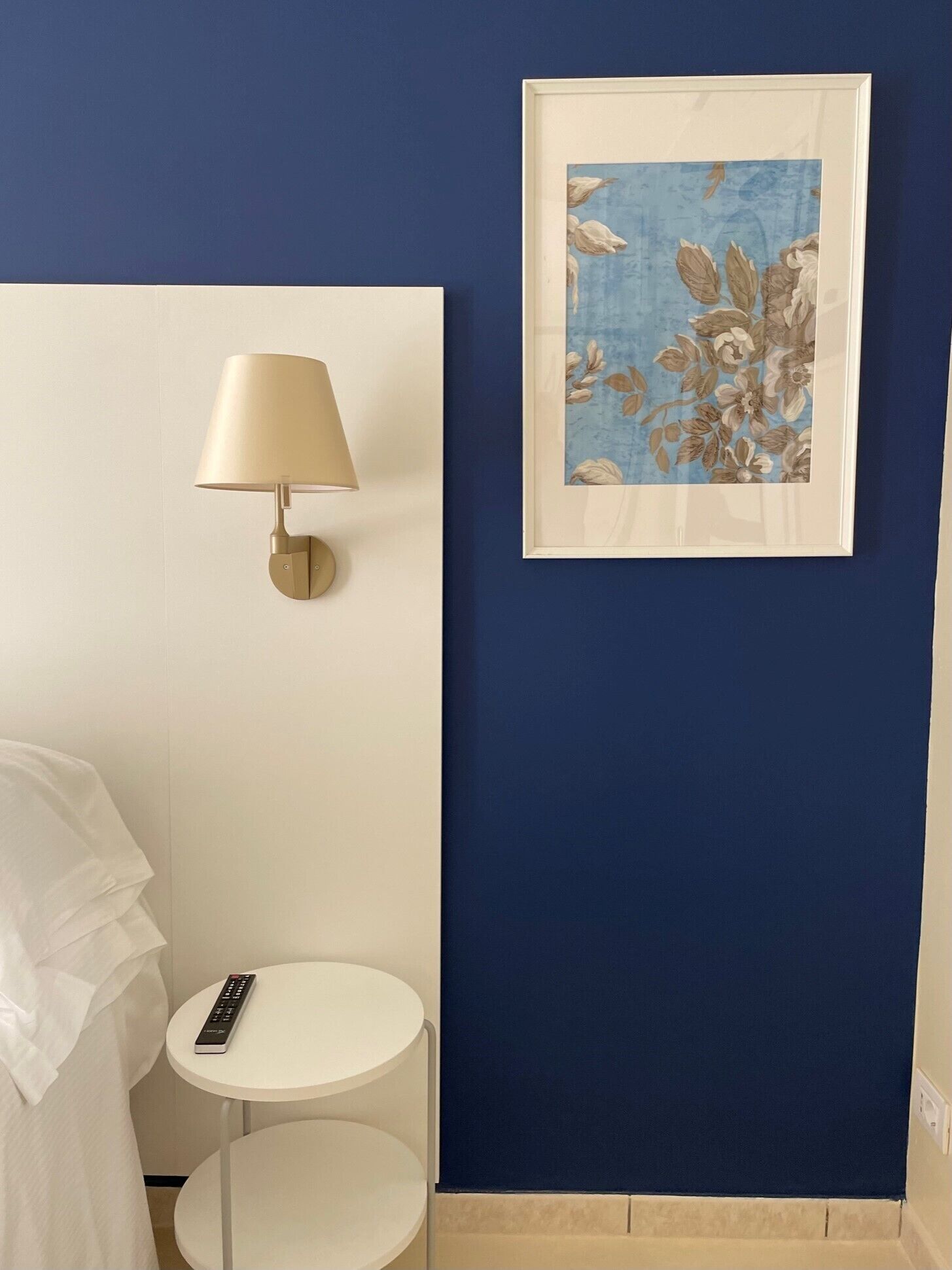 Habitación con pared azul, una obra de arte floral enmarco y una lámpara de pared, junto a una pequeña mesa redonda con un control remoto.