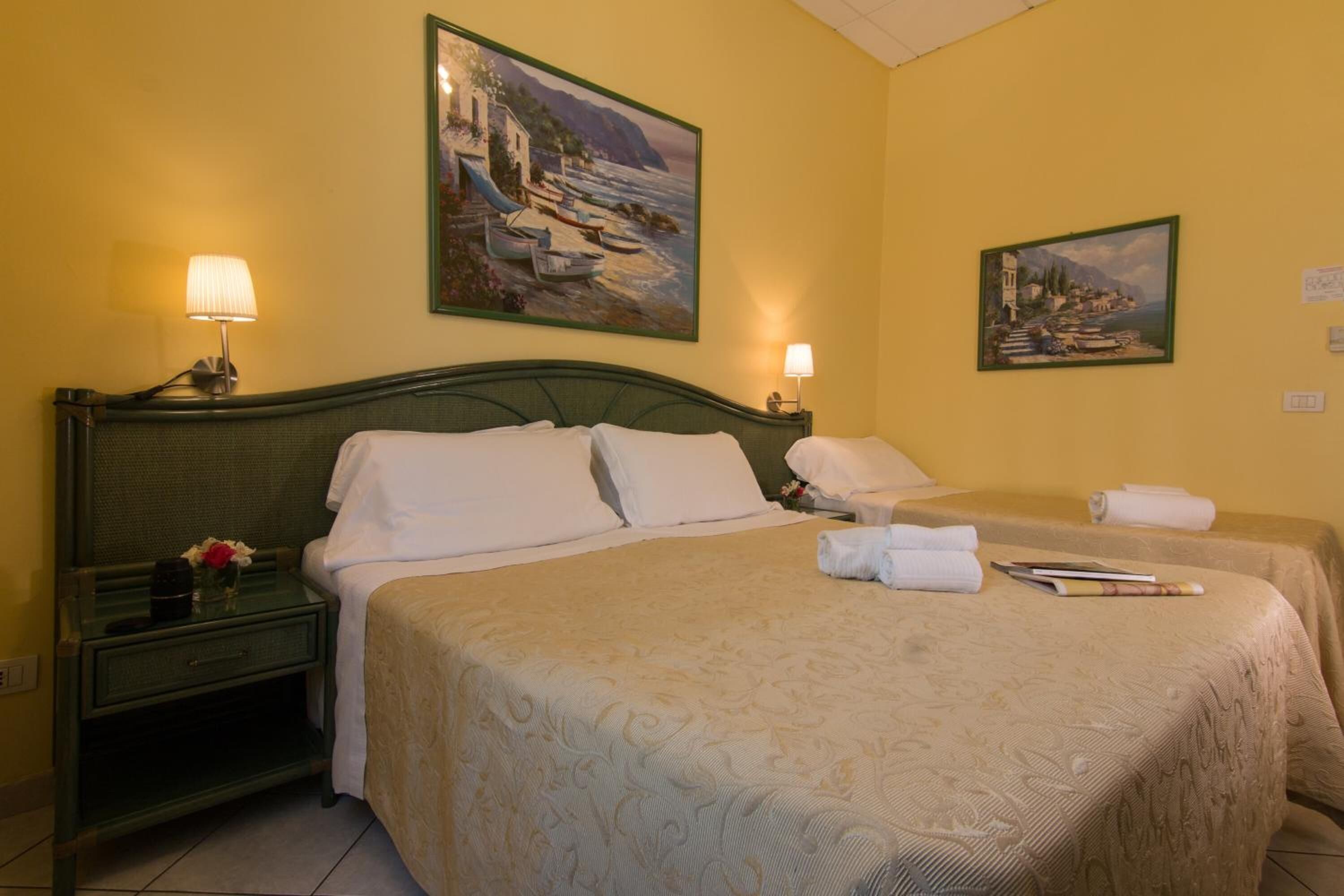 Habitación de hotel con dos camas, una mesita de noche, una lámpara y dos cuadros en la pared.