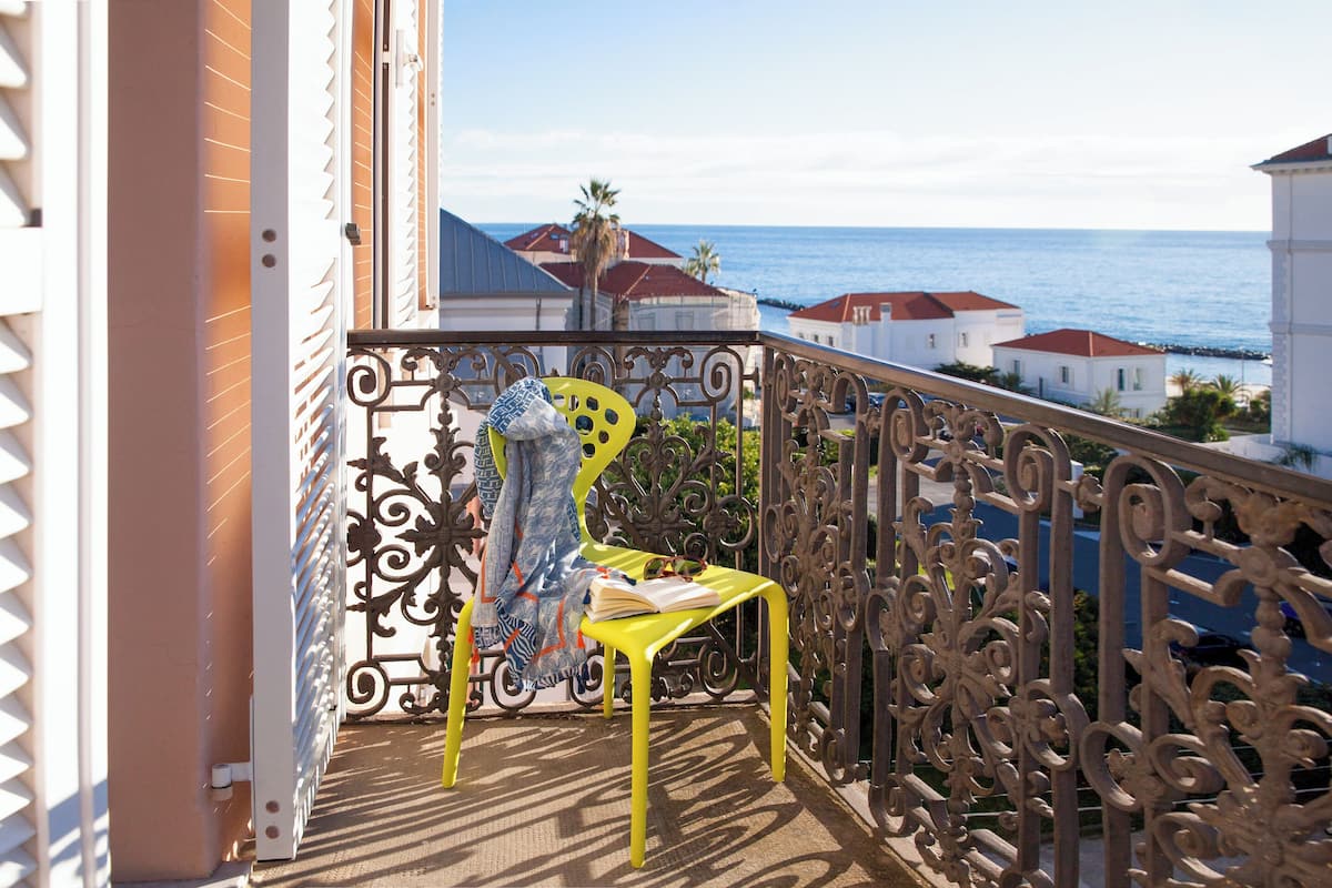 Un balcon avec une chaise jaune, une rambarde en métal décorative et une vue sur la mer et les bâtiments.
