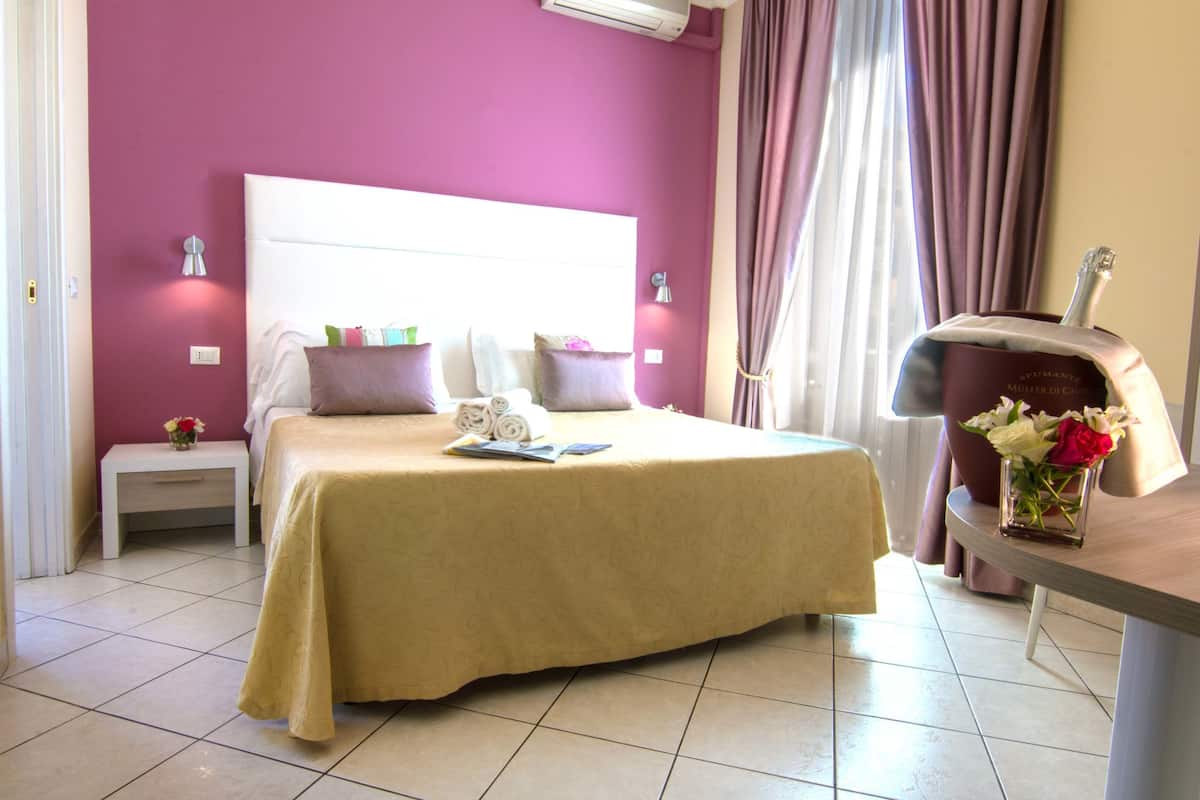 Une chambre d’hôtel avec un lit, des tables de chevet, une petite table, un bureau et un vase de fleurs.