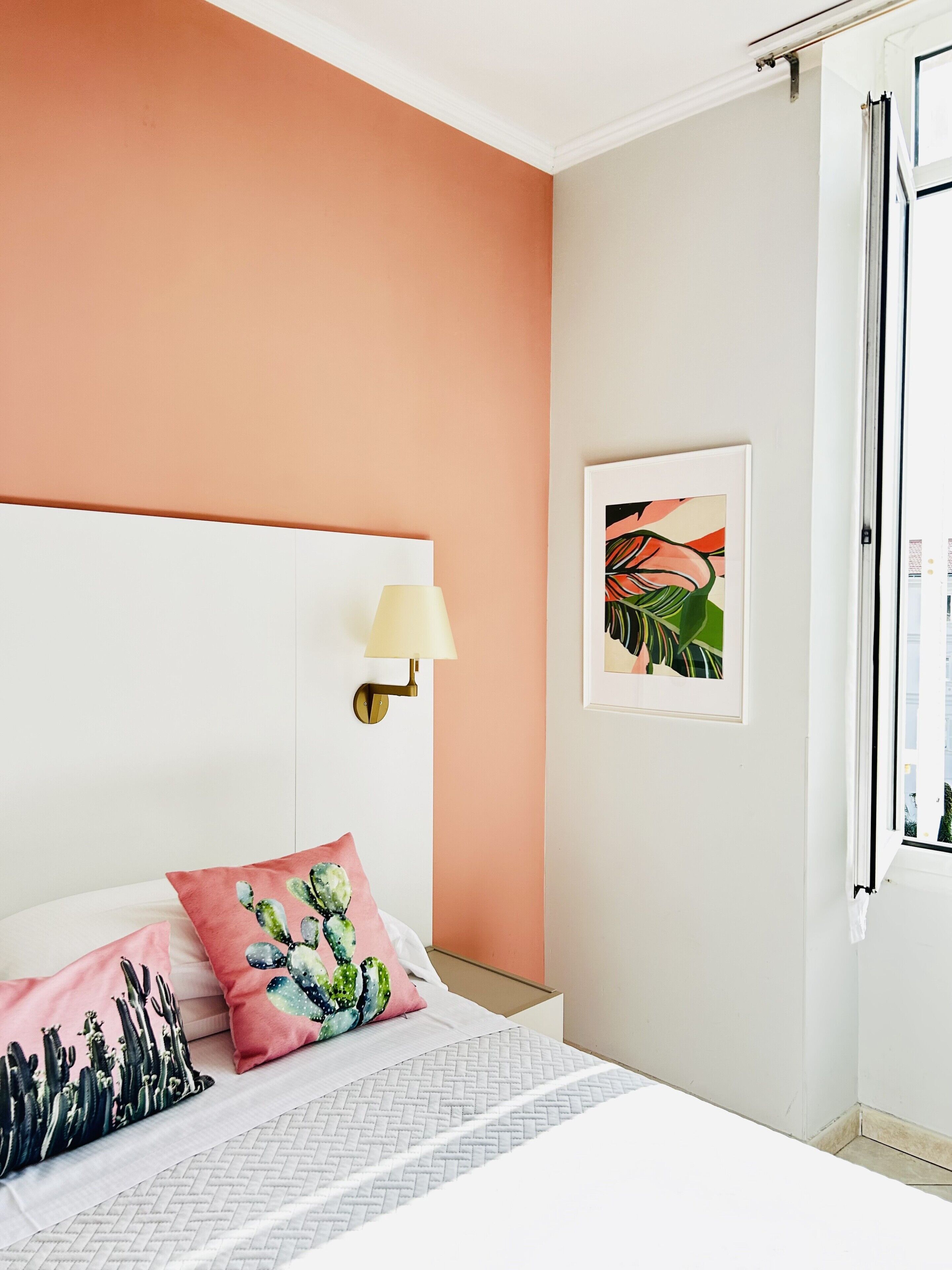 Un dormitorio con cama blanca, paredes color melocotón, almohada con motivos de cactus, lámpara de pared e imagen enmarcada.