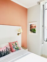 Un dormitorio con cama blanca, paredes color melocotón, almohada con motivos de cactus, lámpara de pared e imagen enmarcada.