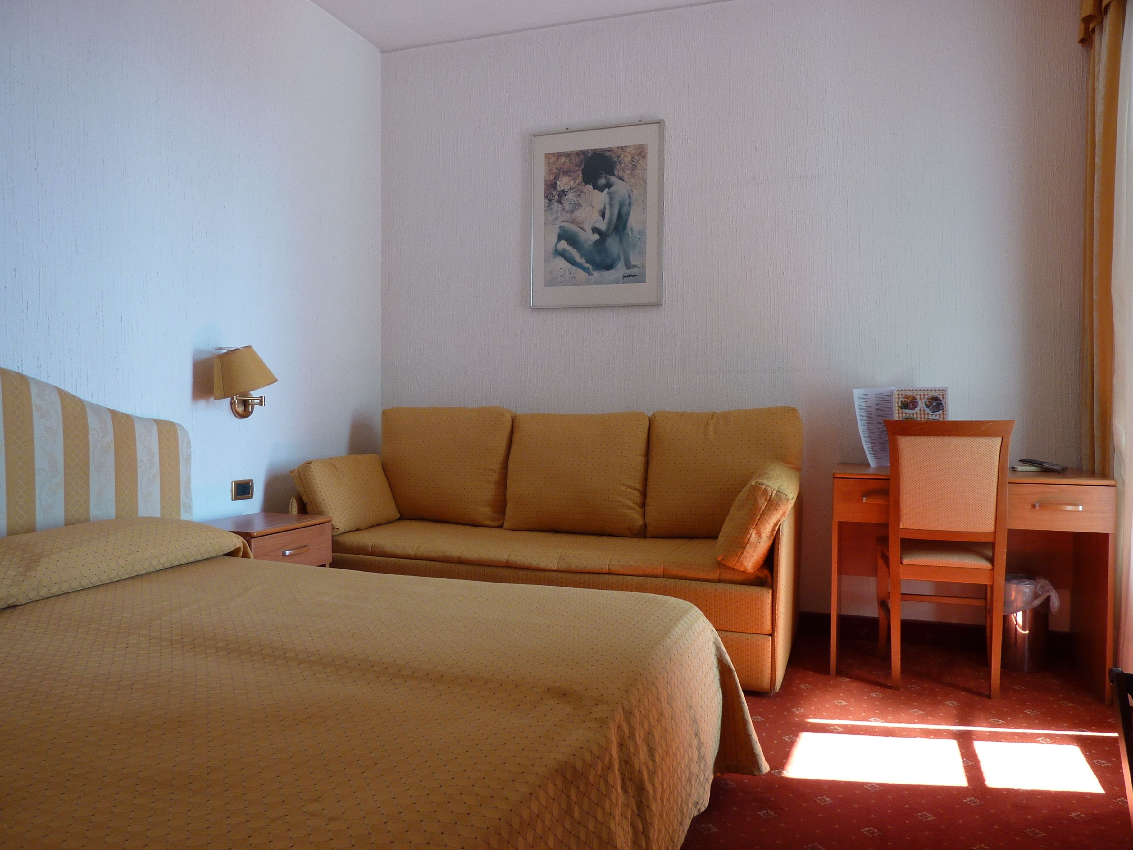 A hotel room with a bed, a sofa, a desk with a chair, a lamp, and a framed picture on the wall.