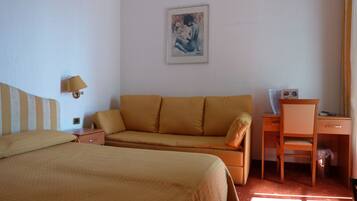 A hotel room with a bed, a sofa, a desk with a chair, a lamp, and a framed picture on the wall.