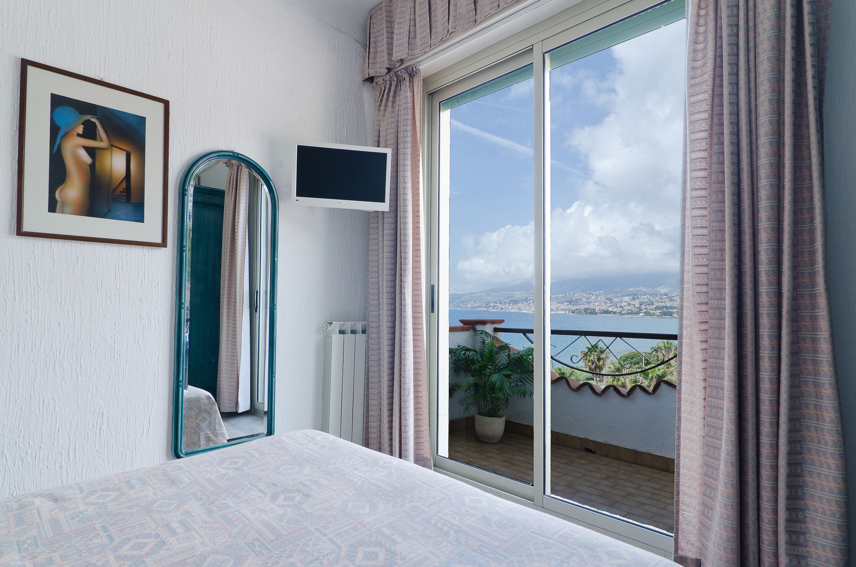 Un dormitorio con un ventanal grande, una cama, un espejo, un televisor y un cuadro en la pared.