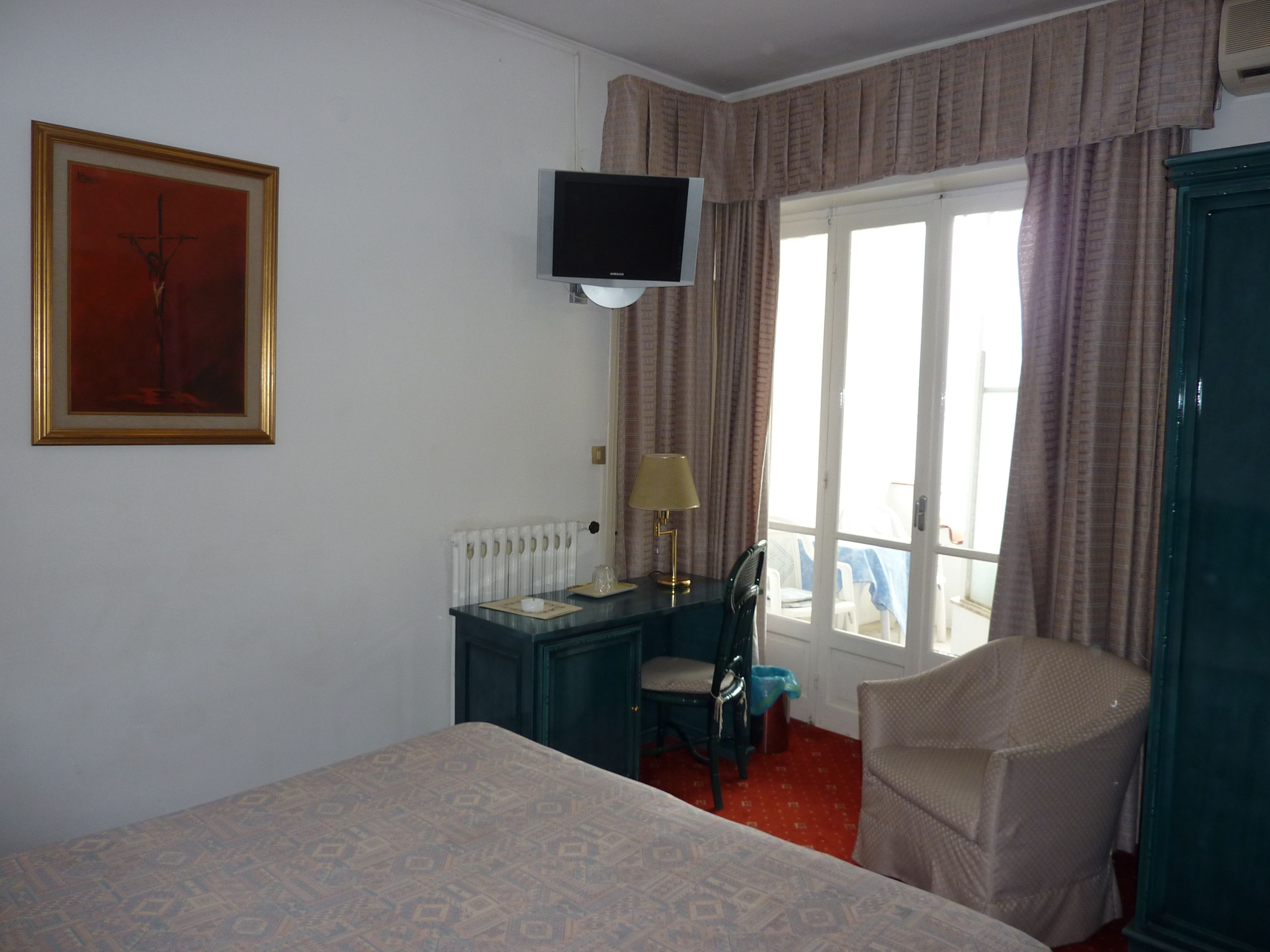 A hotel room with a bed, a desk with a chair, a television on the wall, and a window with curtains.
