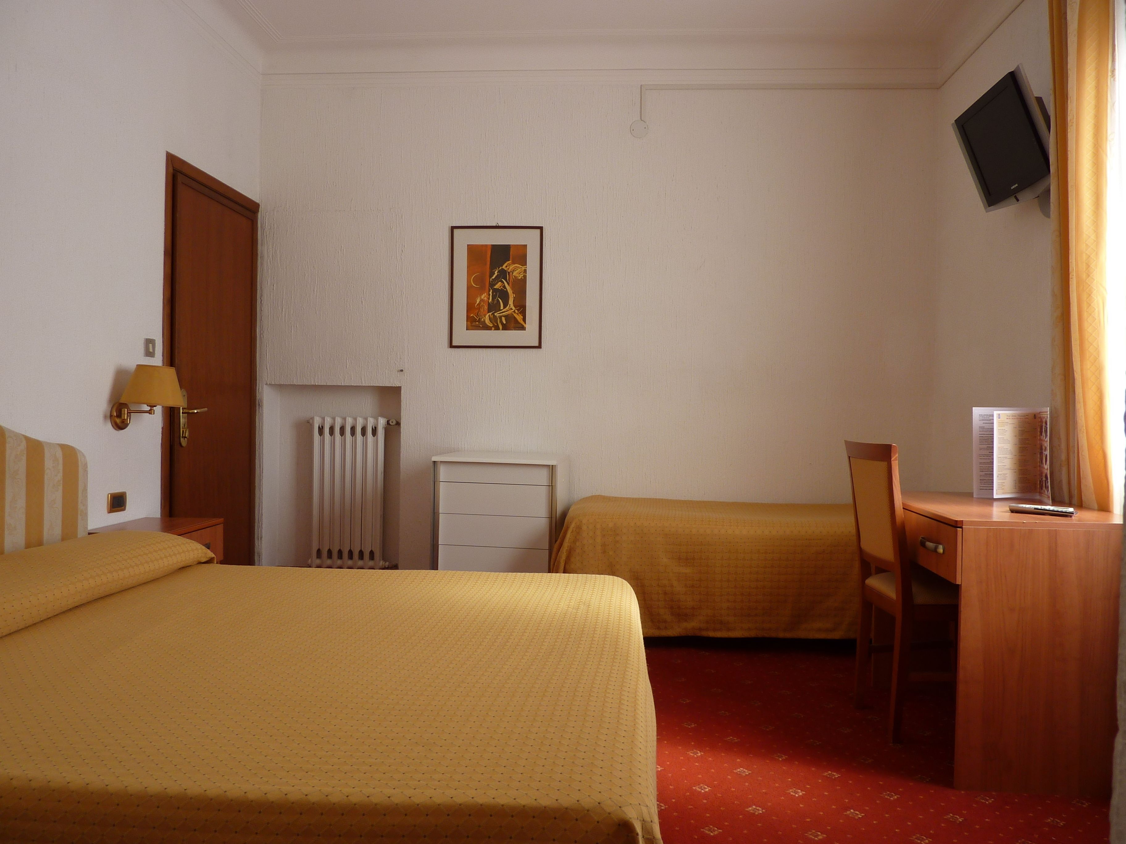 A hotel room with two beds, a desk, a chair, a radiator, a television, and a framed picture on the wall.