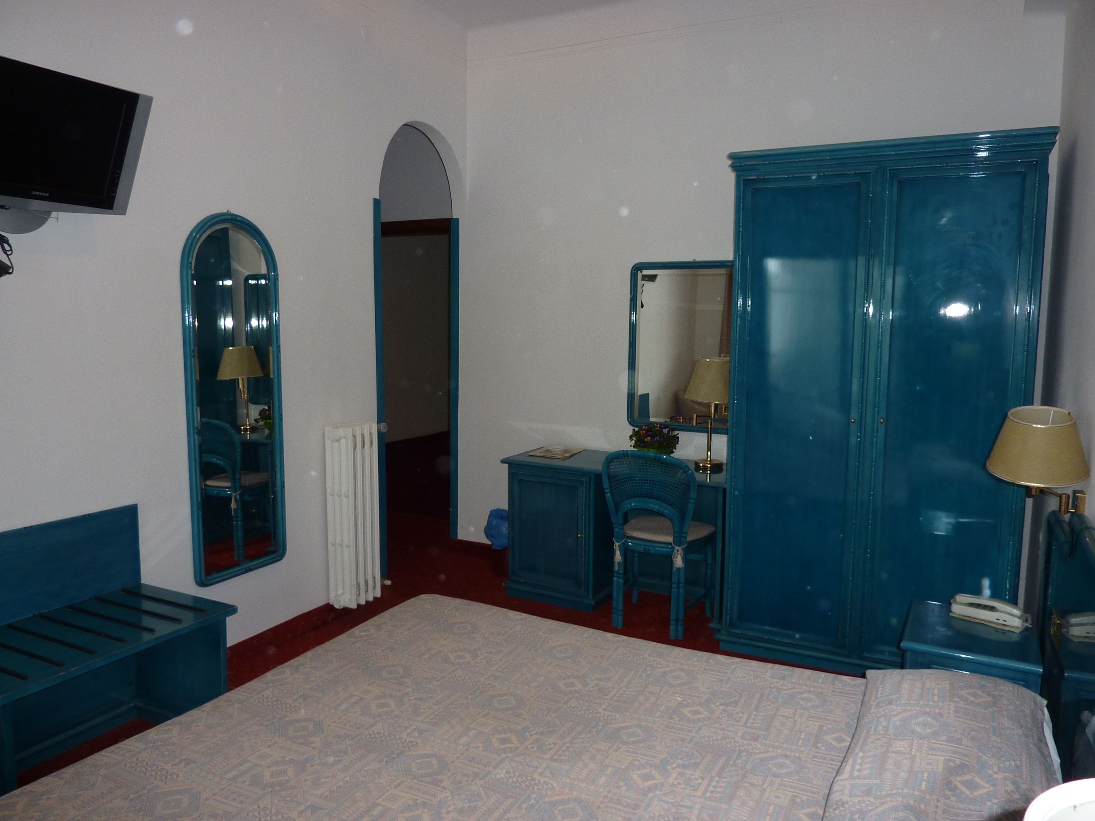 A bedroom with a bed, a dresser, a desk, a chair, a mirror, a radiator, and a television.