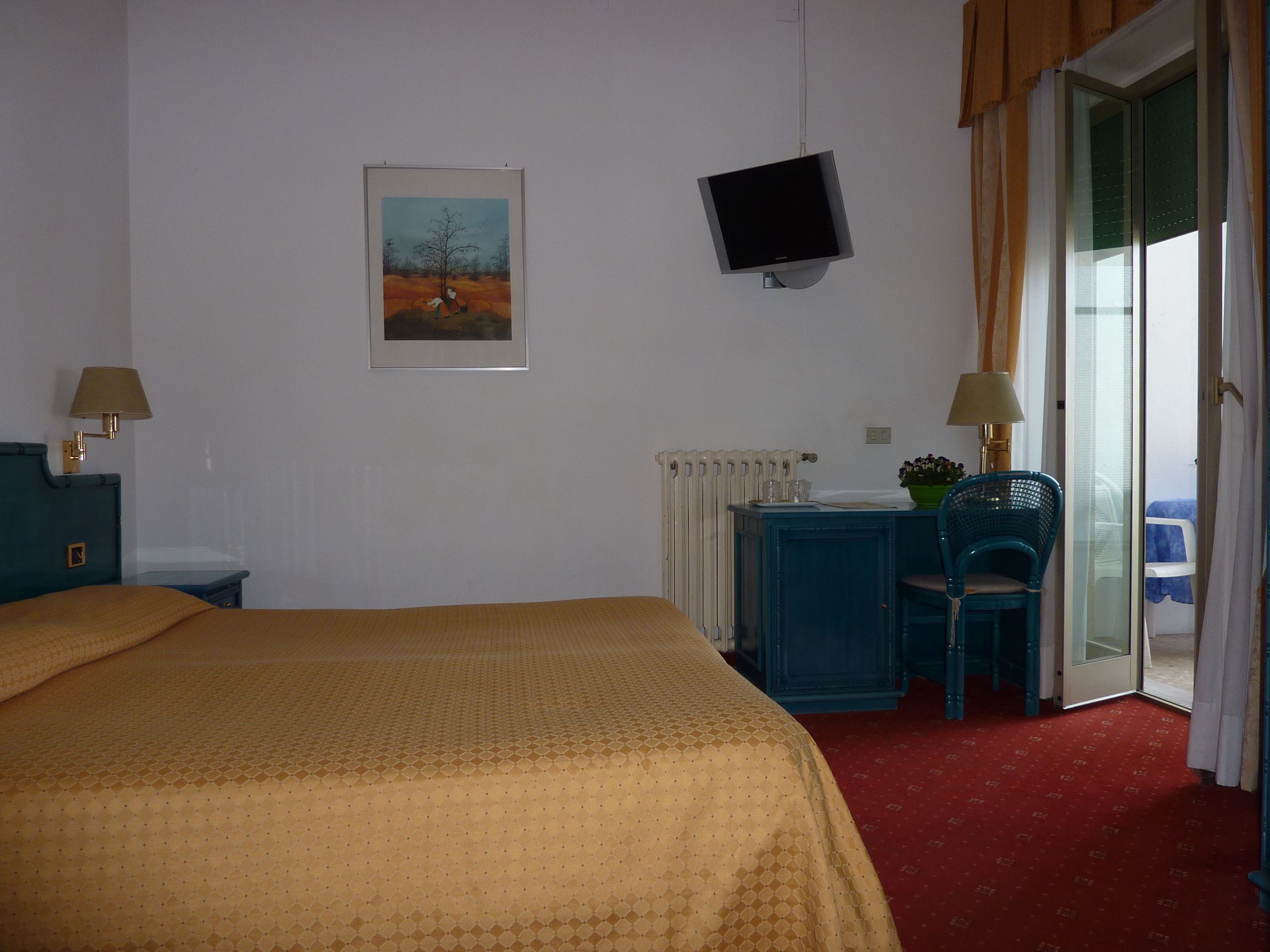 A hotel room with a bed, a desk with a chair, a television, a radiator, and a window with curtains.