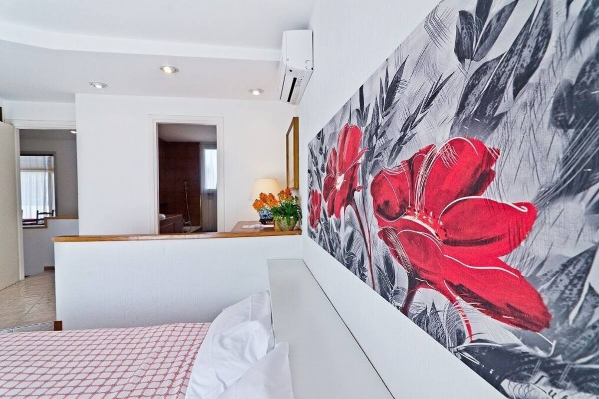 Een moderne hotelkamer met een groot bloemenkunstwerk aan de muur, een bed met een geborduurde spreideken, en een bureau met een lamp en bloemen.