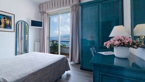 Een slaapkamer met een bed, een televisie, een raam met gordijnen, uitzicht op zee, en een blauwgroene ladekast met een lamp en bloemen.