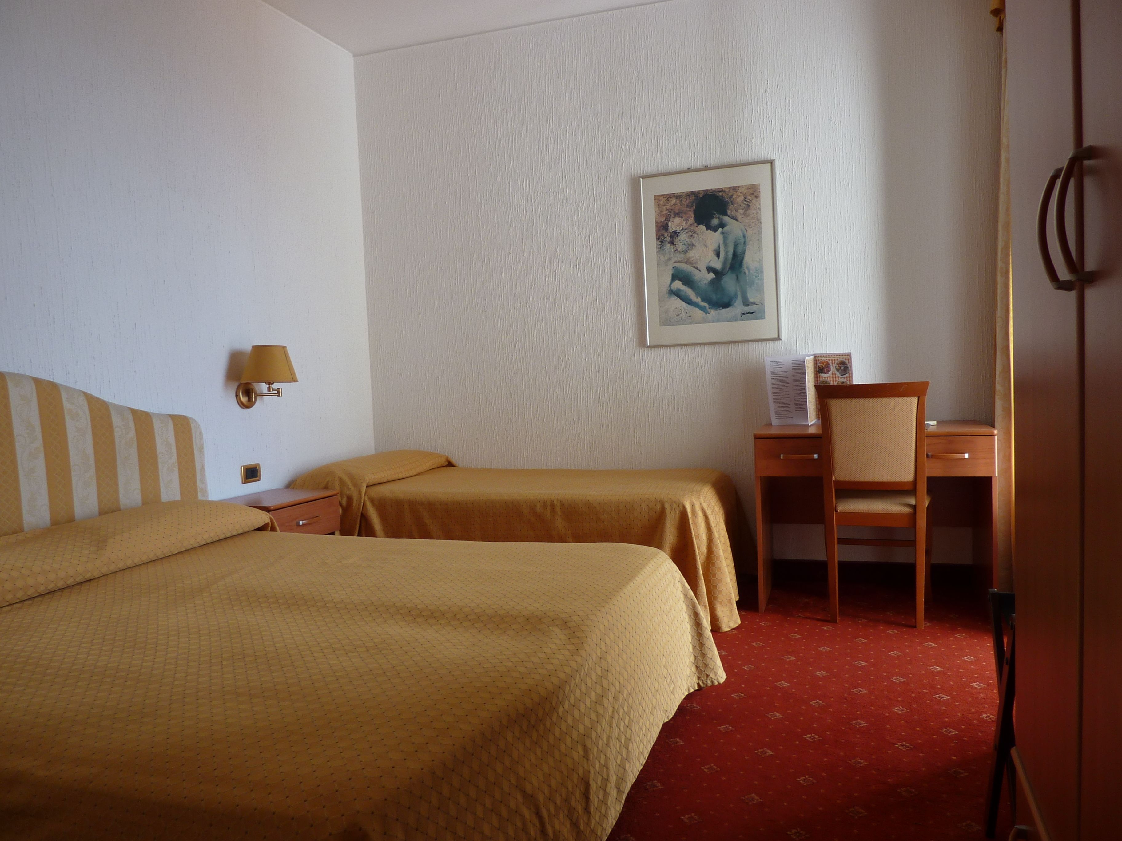 Habitación de hotel con dos camas individuales, un escritorio, una silla, una lámpara y un cuadro en la pared.