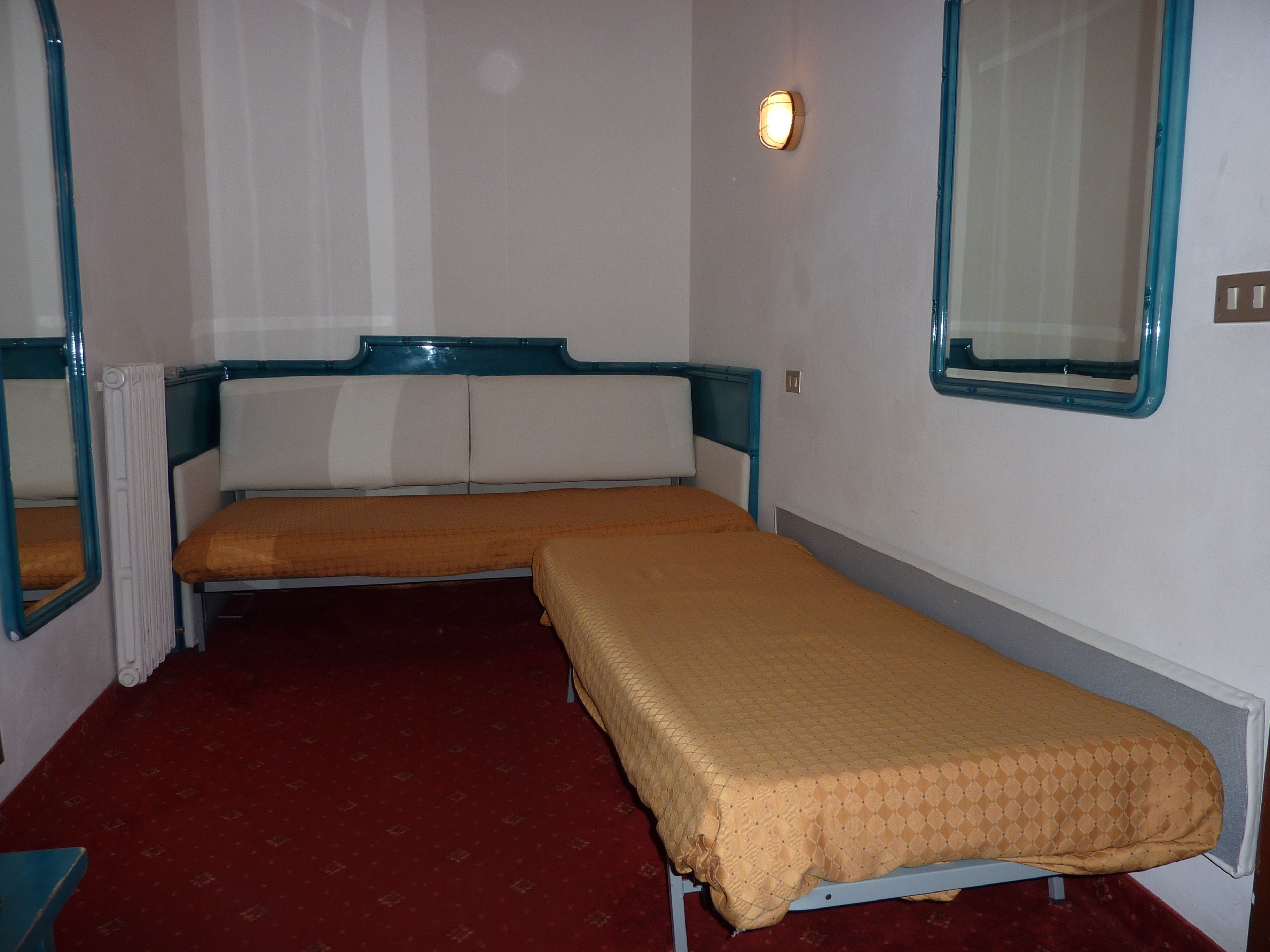 A small, single-bed room with a red carpet, a window with a blue frame, and a wall-mounted light fixture.