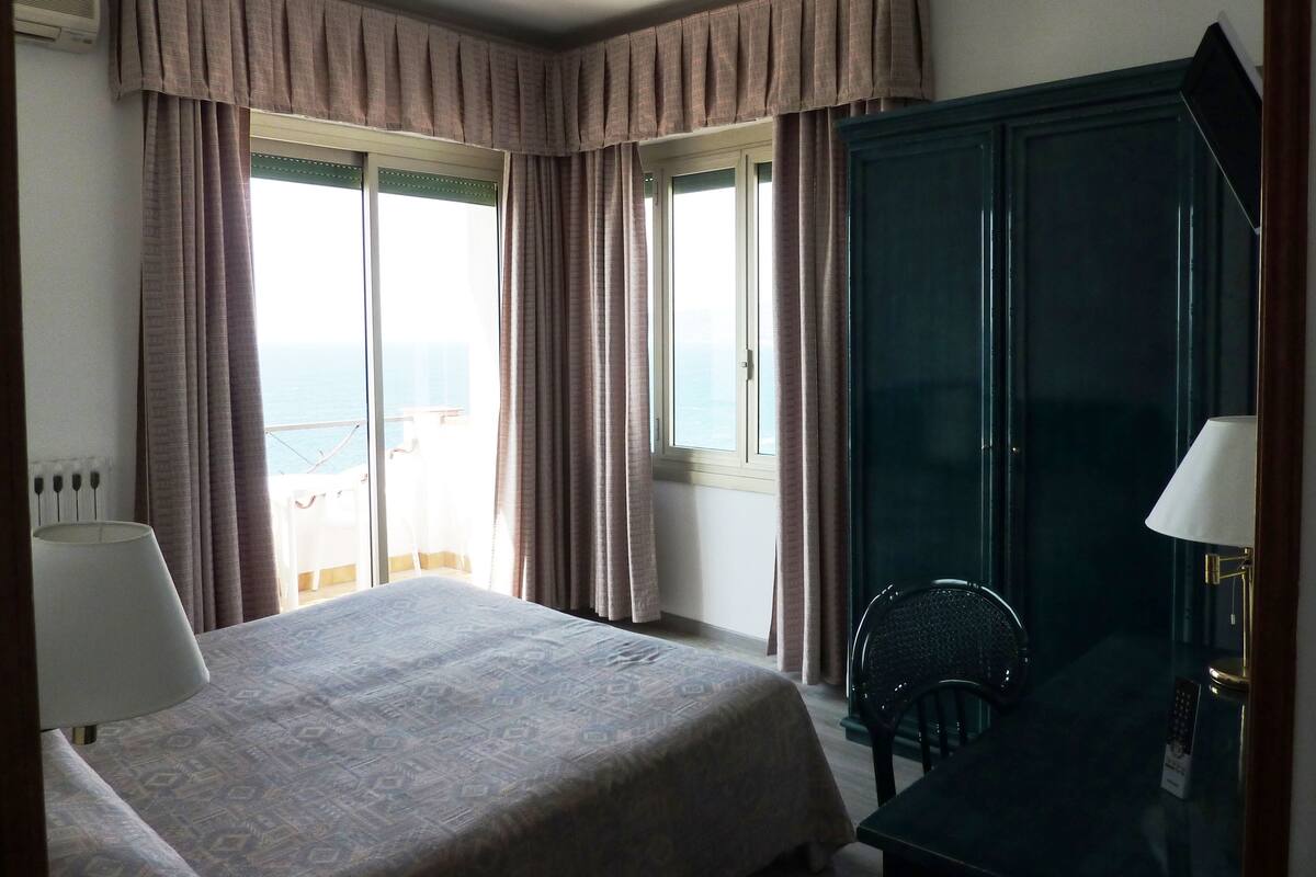 Een hotelkamer met een bed, een bureau, een stoel en uitzicht op zee door grote ramen.
