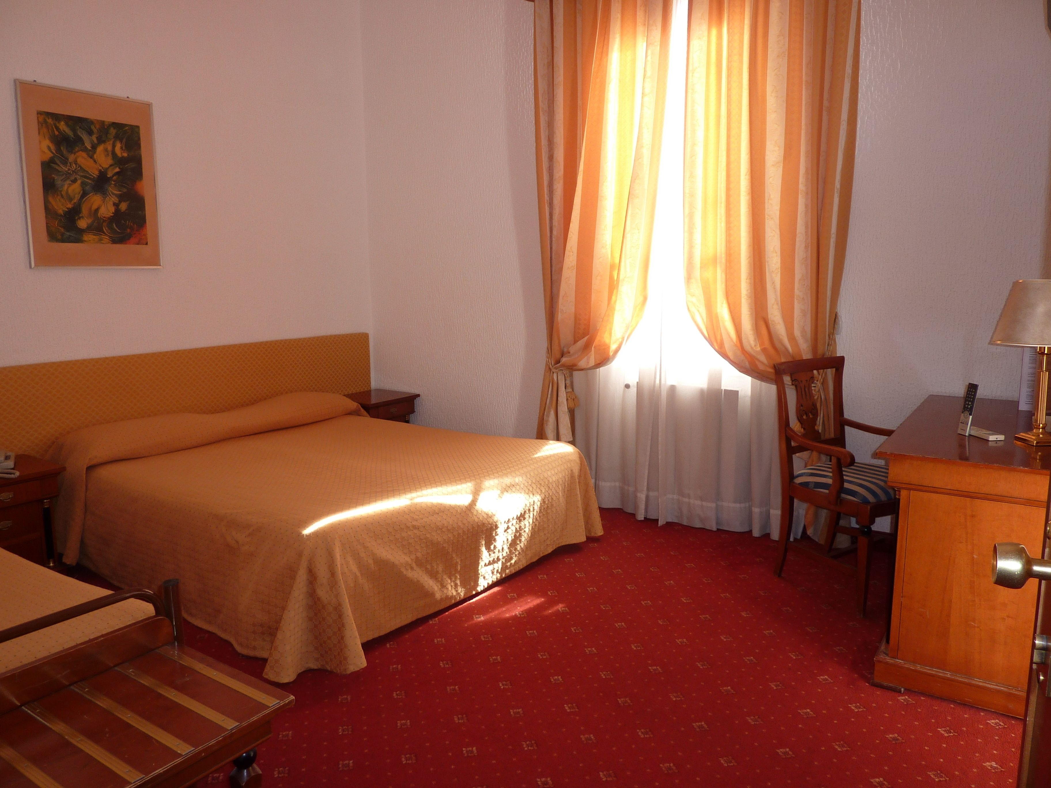 A hotel room with a bed, a desk with a lamp, a chair, and a painting on the wall.