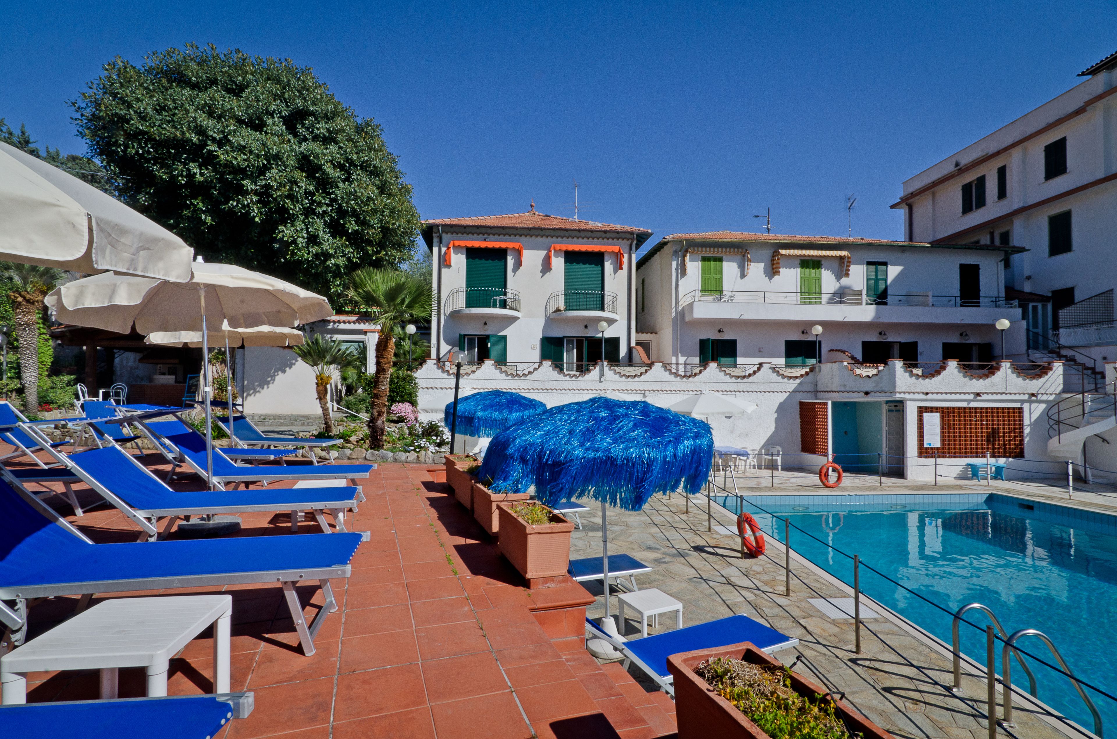 Un área junto a la piscina con sillones y sombrillas, una piscina y edificios al fondo.