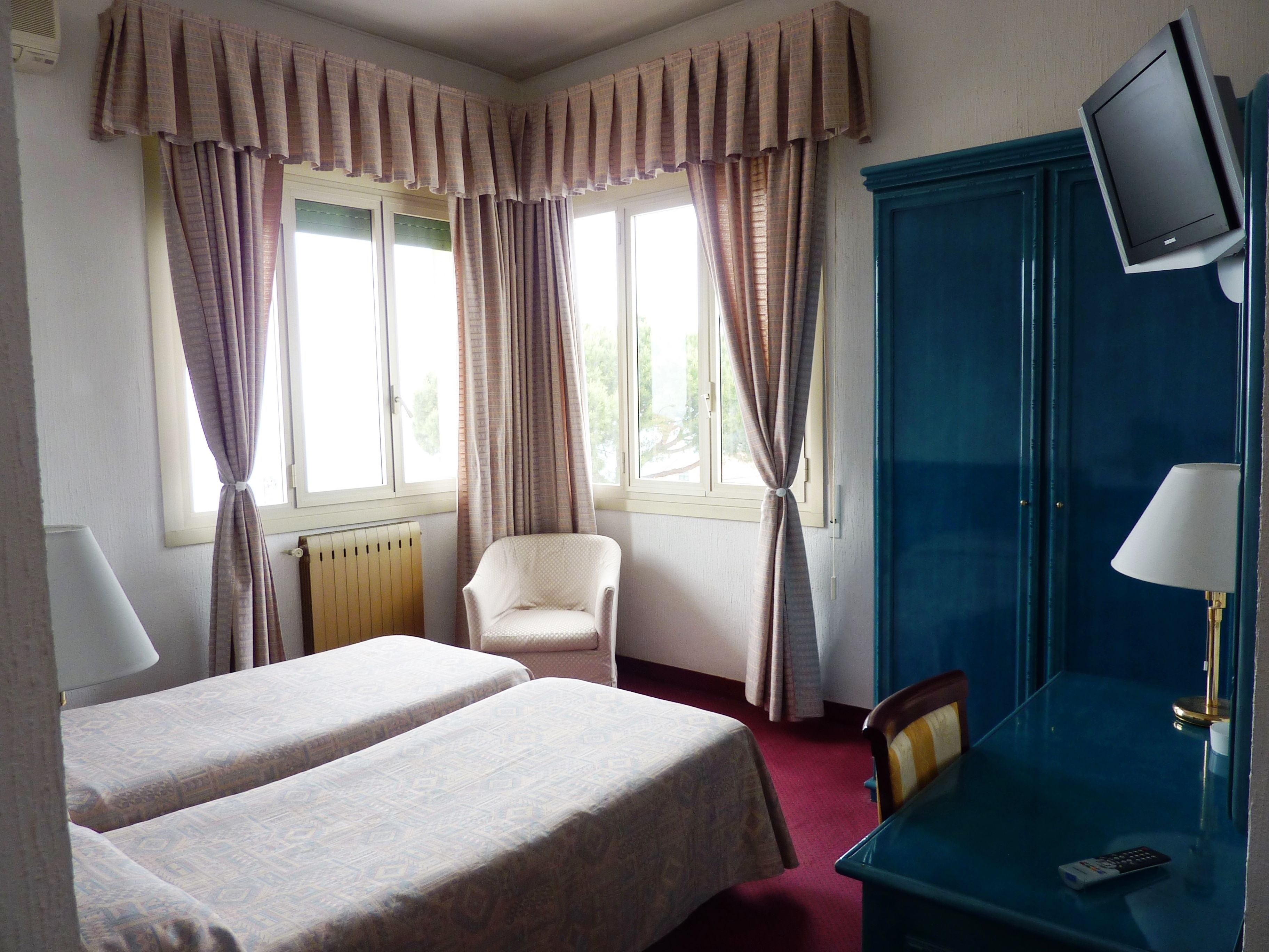 Ein Hotelzimmer mit zwei Betten, einem Sessel, einem Schreibtisch mit Lampe, einem Fernseher und einem Fenster mit Vorhängen.
