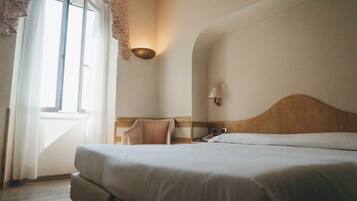 Ein Hotelzimmer mit einem großen Bett, einem hölzernen Kopfteil, einem Nachttisch mit Lampe und einem Fenster mit leichtem Vorhang.