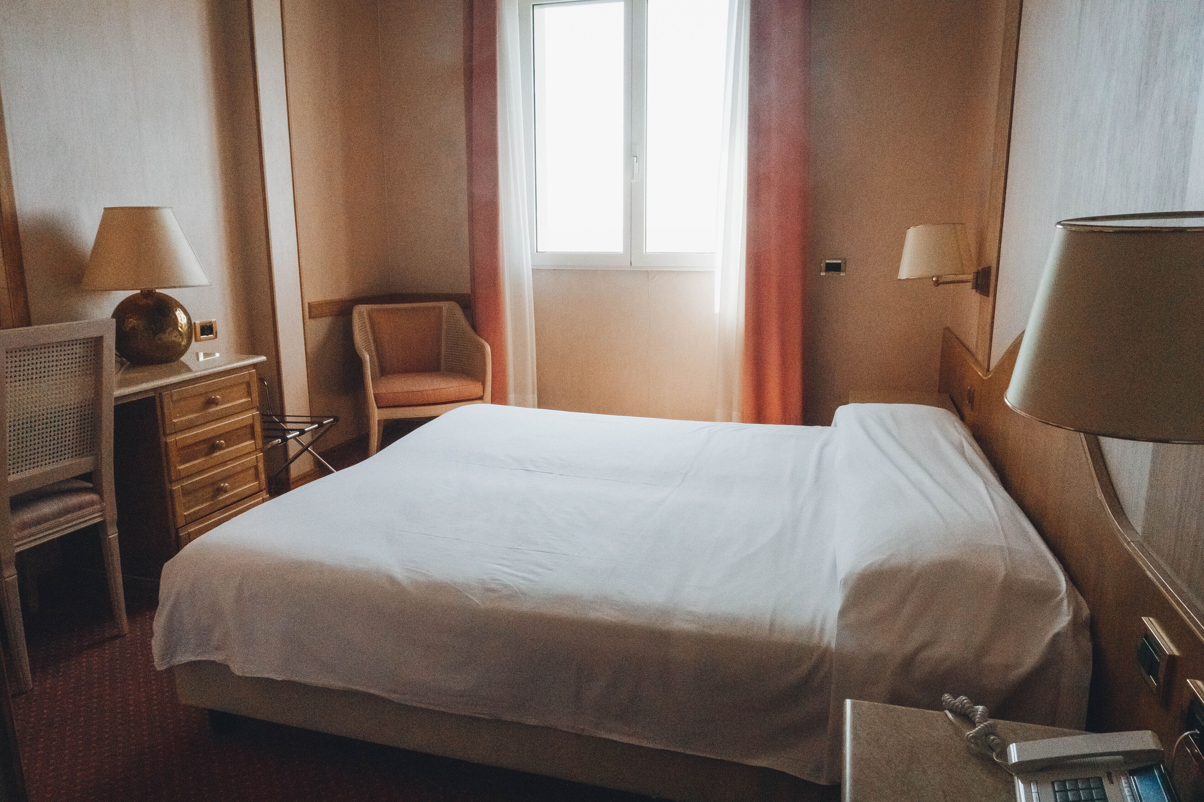 Ein Hotelzimmer mit Bett, Sessel, Schreibtisch mit Lampe und Fenster mit Vorhängen.