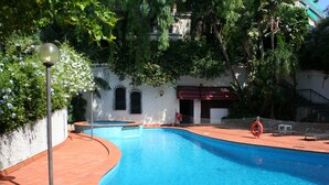 Una piscina con borde curvo, rodeada por un borde de azulejos y un muro blanco con un toldo rojo.