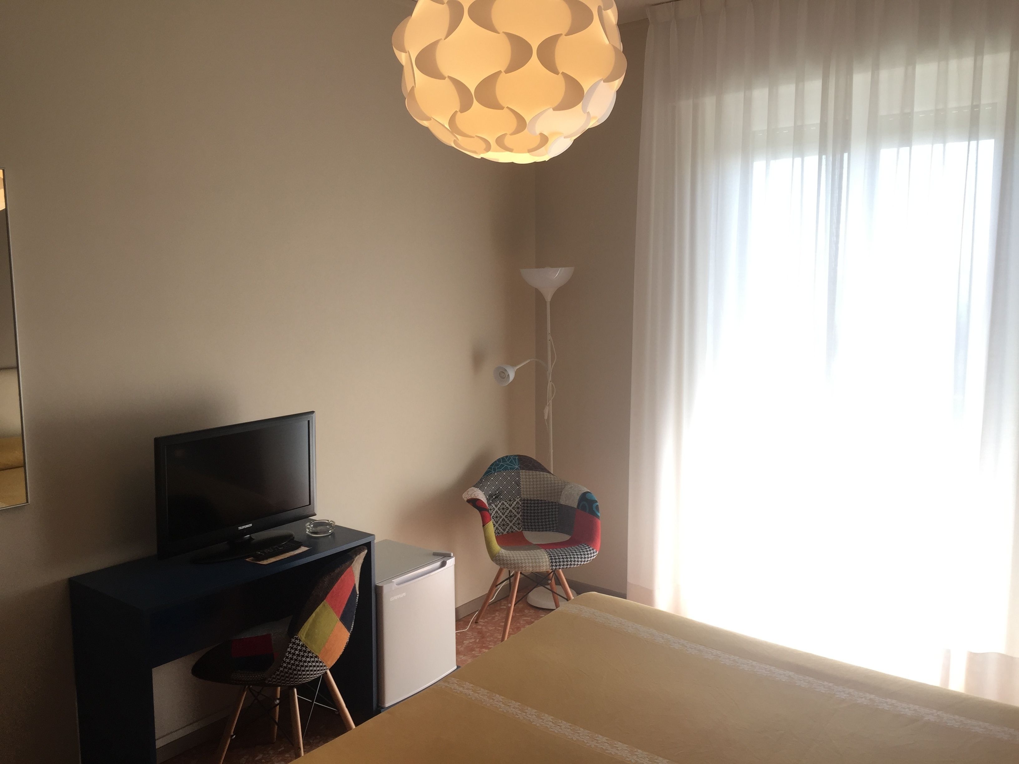 Habitación de hotel con cama, escritorio con televisor, una silla, lámpara y ventana con cortinas.