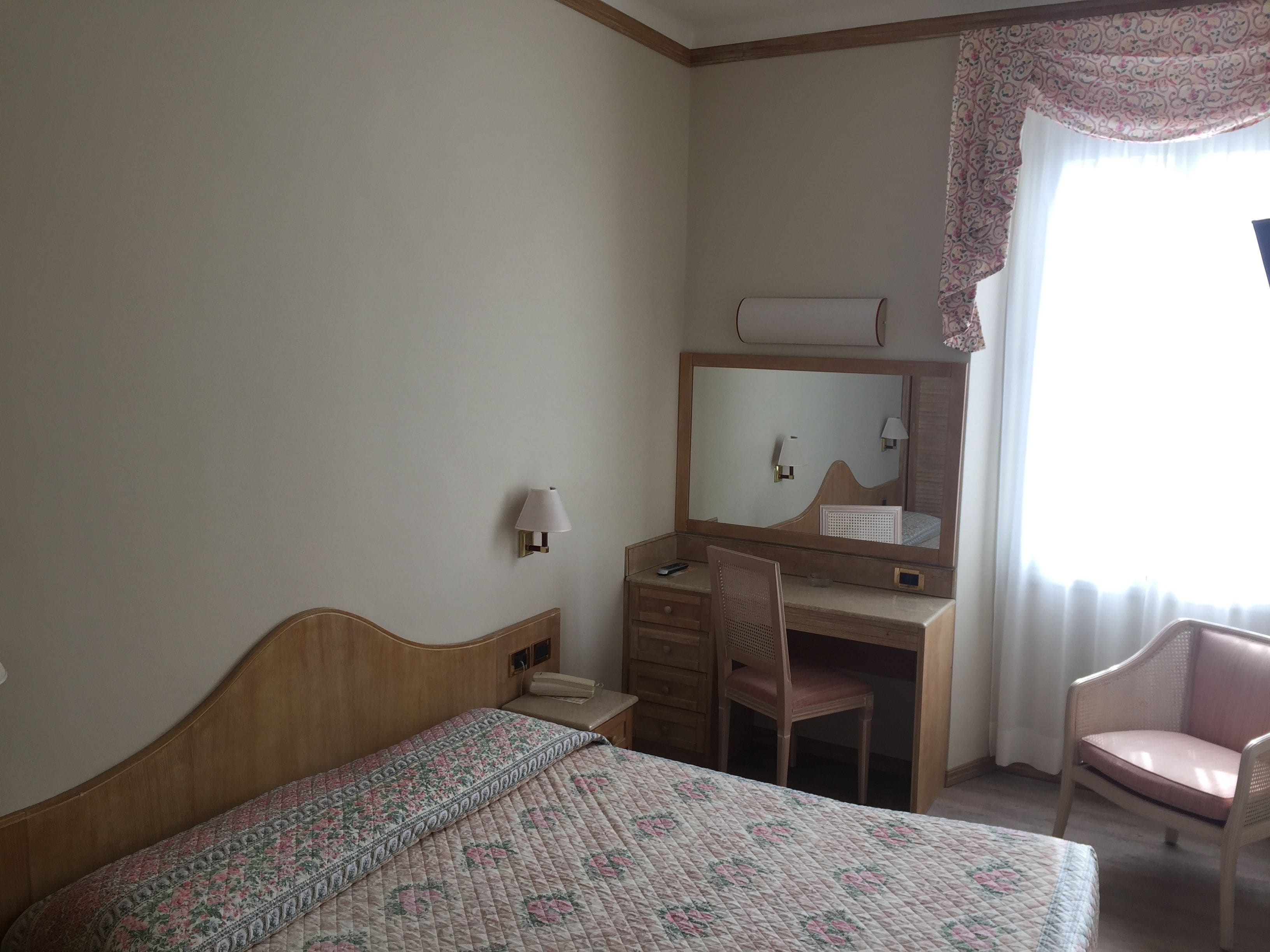 A hotel room with a bed, a desk, a chair, a mirror, and a window with curtains.