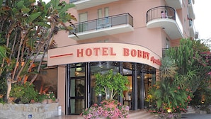 Entrada de un hotel con un letrero curvo, puertas de vidrio y un pequeño jardín con flores.
