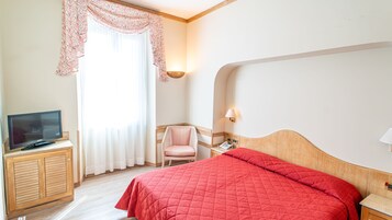 A hotel room with a bed, a TV on a wooden cabinet, a chair, a bedside table with a lamp, and a window with curtains.