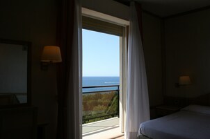 A hotel room with a bed, a nightstand, a lamp, and a view of the sea through a window.