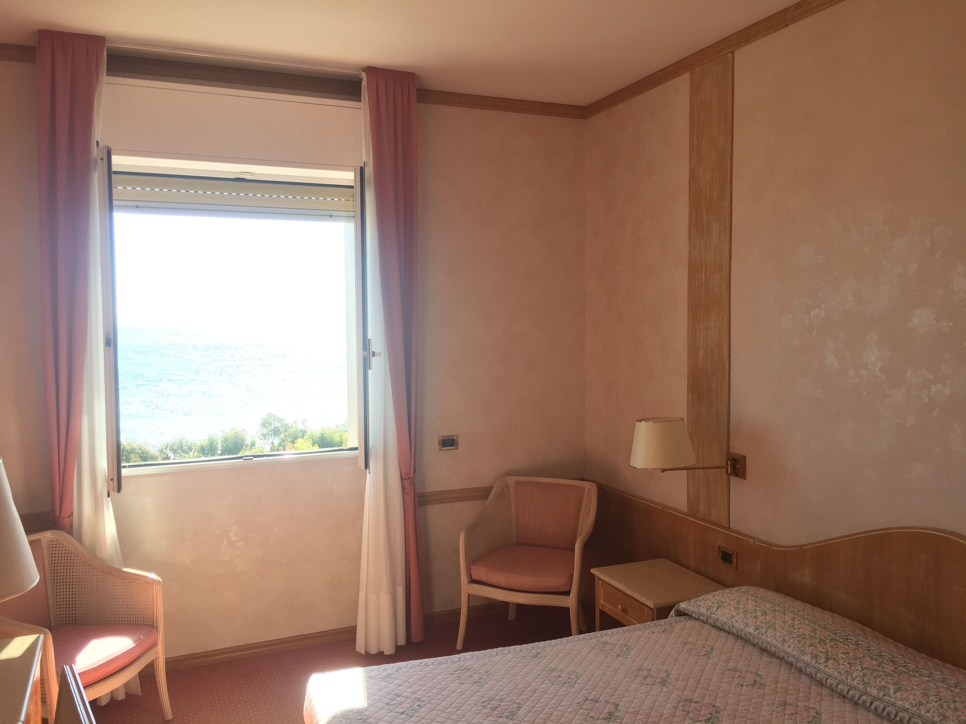 Une chambre d’hôtel avec un lit, une chaise, une table de chevet, une lampe et une fenêtre avec des rideaux.