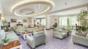 A spacious living room with multiple sofas and armchairs arranged around a central coffee table. There are several potted plants and a television on a wooden stand. The room has a patterned carpet and large windows with green curtains. - Piccolo Paradiso (Massa Lubrense)