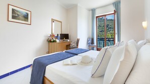 A hotel room with a bed, a desk with a computer, a chair, and a window with a view of greenery. - Piccolo Paradiso (Massa Lubrense)