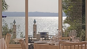 A dining area with a table set for two, overlooking a body of water with a distant shoreline. - Hotel Marconi (Sirmione)