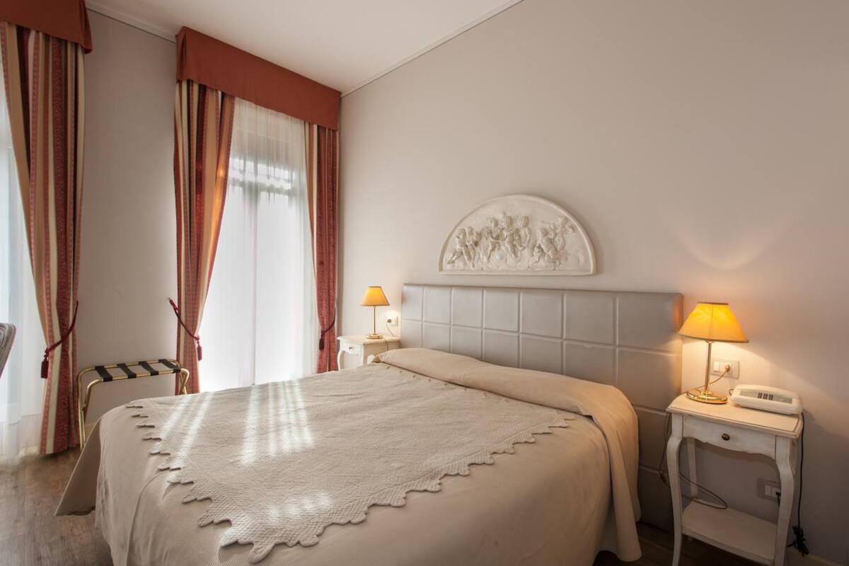 A hotel room with a bed, two bedside lamps, a nightstand with a phone, a chair, and a window with curtains.