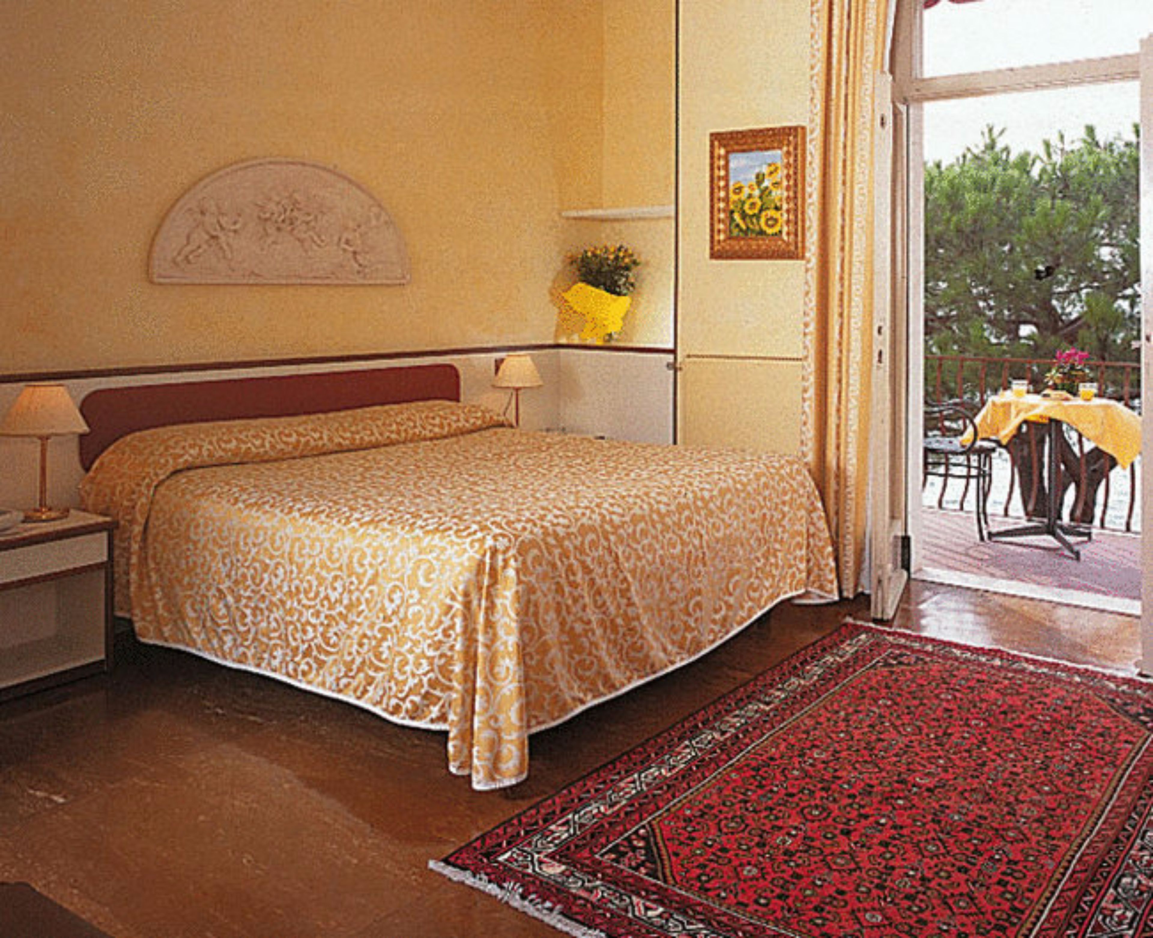 A bedroom with a bed, bedside tables, a nightstand, a window with curtains, a framed picture on the wall, and a view of the outdoors.