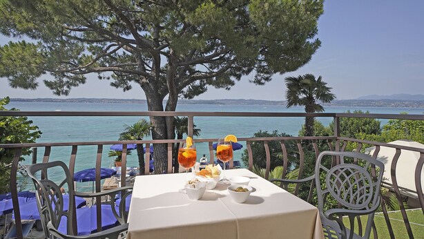 A table set with drinks and snacks, overlooking a body of water with a tree in the background. - Hotel Marconi (Sirmione)