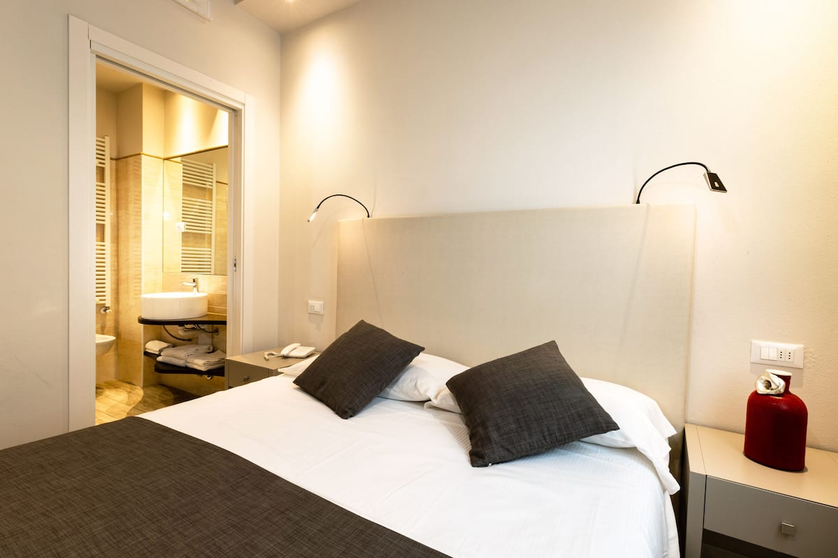 A hotel room with a bed, bedside table, nightstand, and a bathroom visible through an open door.