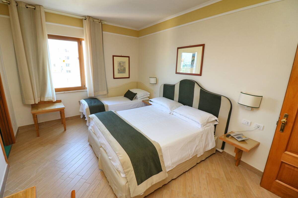 A hotel room with two beds, a small table, a bedside table with a phone, and a window with curtains.