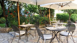 Un patio al aire libre con suelo de piedra, mesas y sillas de metal, rodeado de exuberante follaje y plantas con flores.