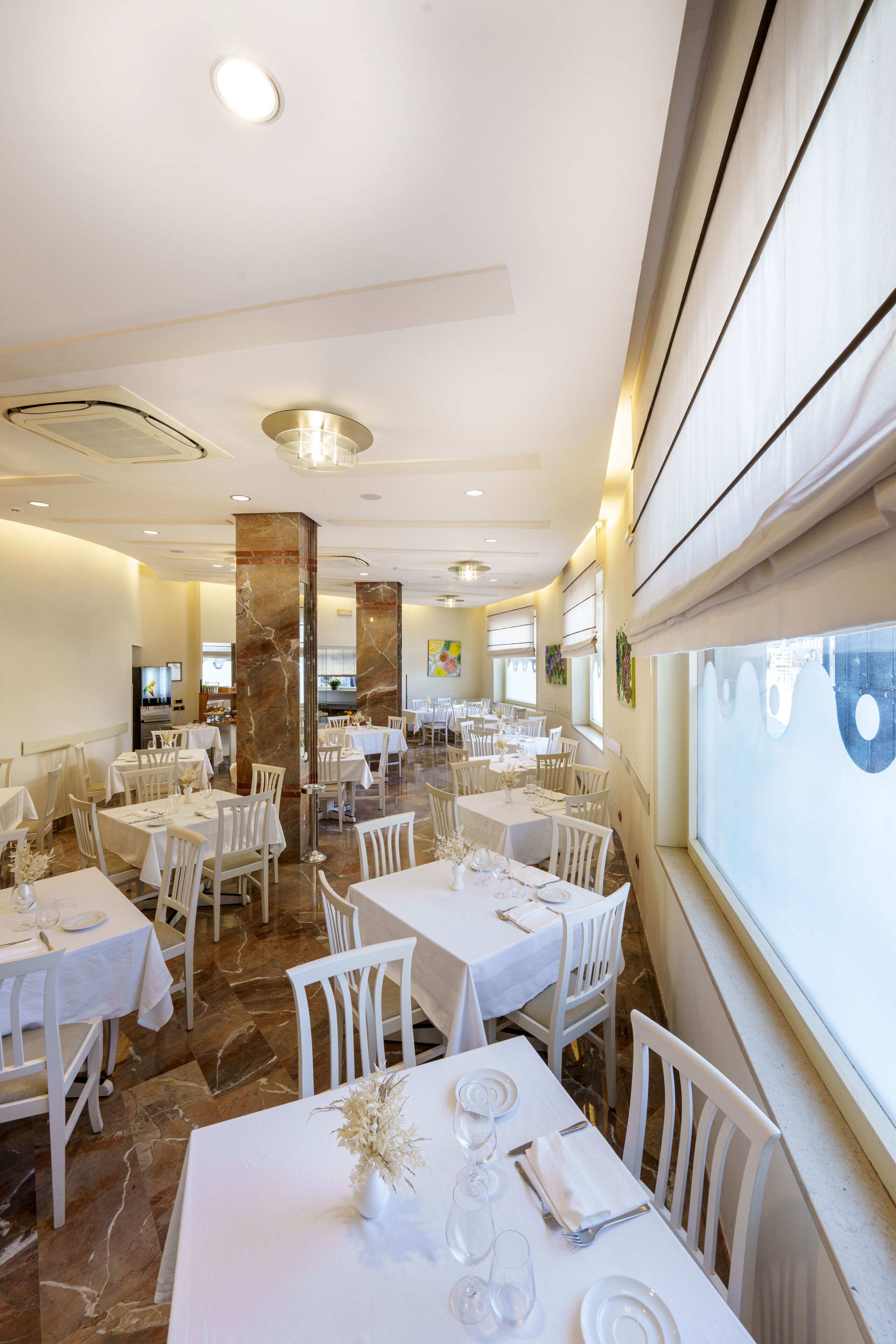 A spacious restaurant with multiple tables and chairs, marble flooring, and large windows.