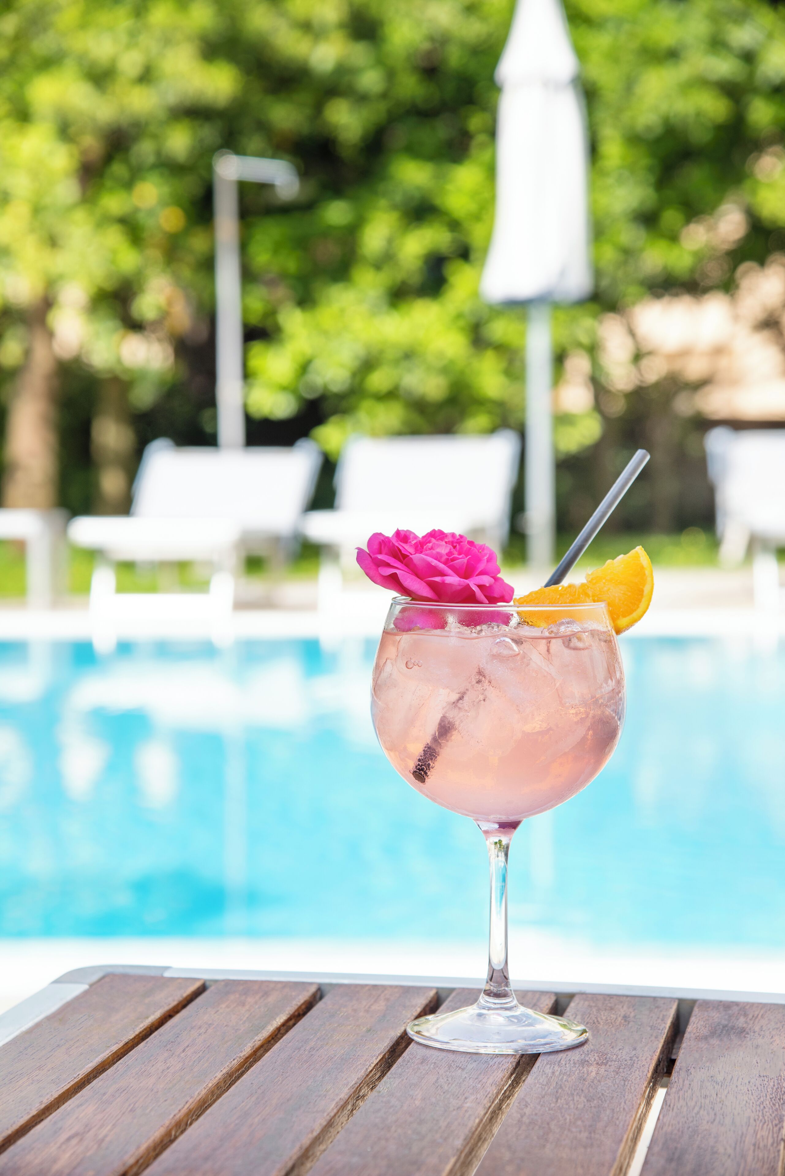 Coquetel com pétala de rosa e fatia de laranja sobre mesa de madeira junto a uma piscina.