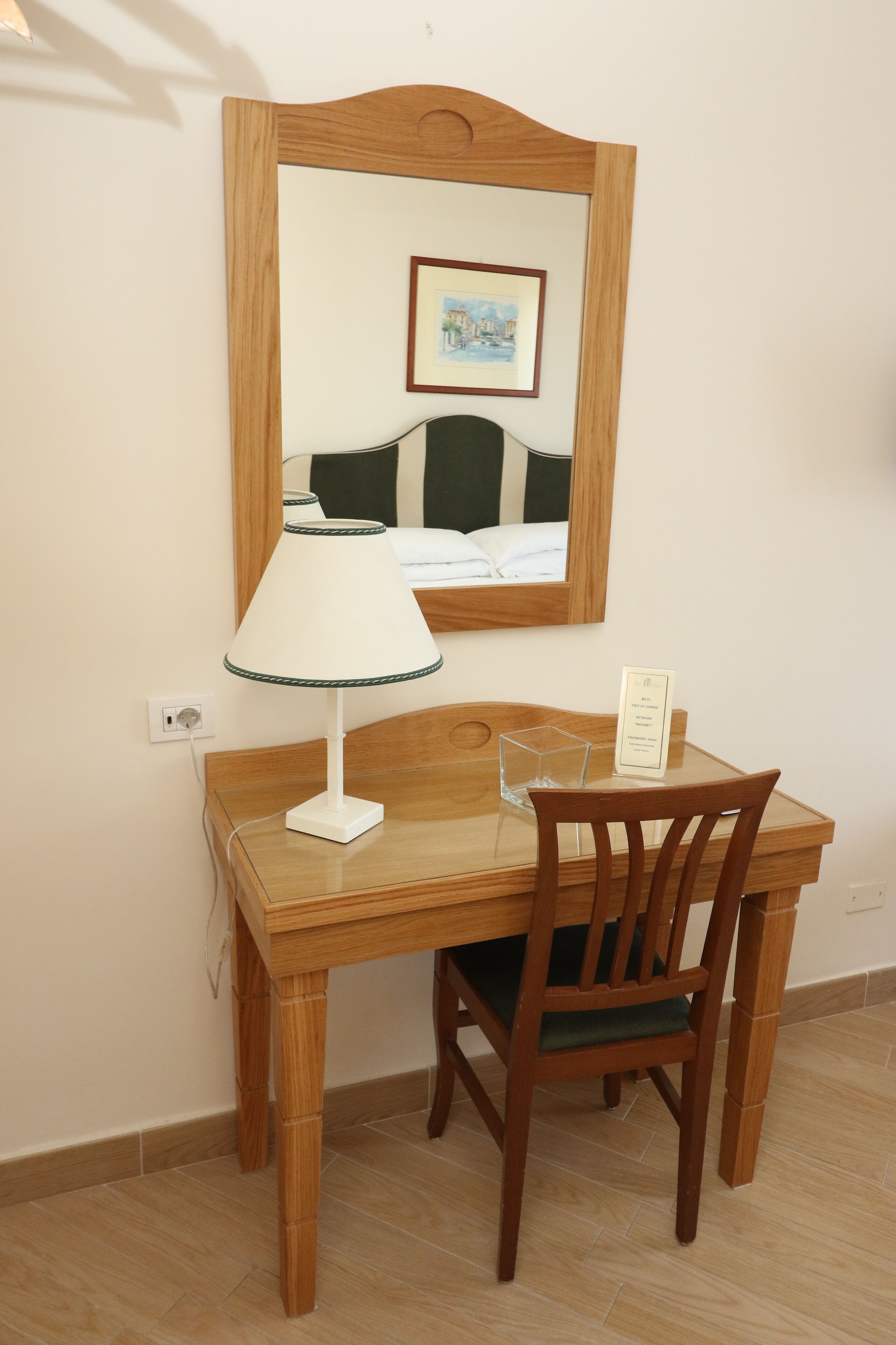 A wooden desk with a lamp, a chair, and a mirror reflecting a bed.