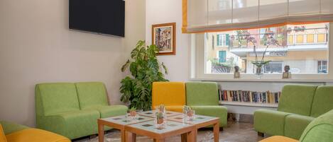 Un salon avec des meubles verts et jaunes, un téléviseur à écran plat, une grande fenêtre donnant sur des immeubles et une plante en pot.