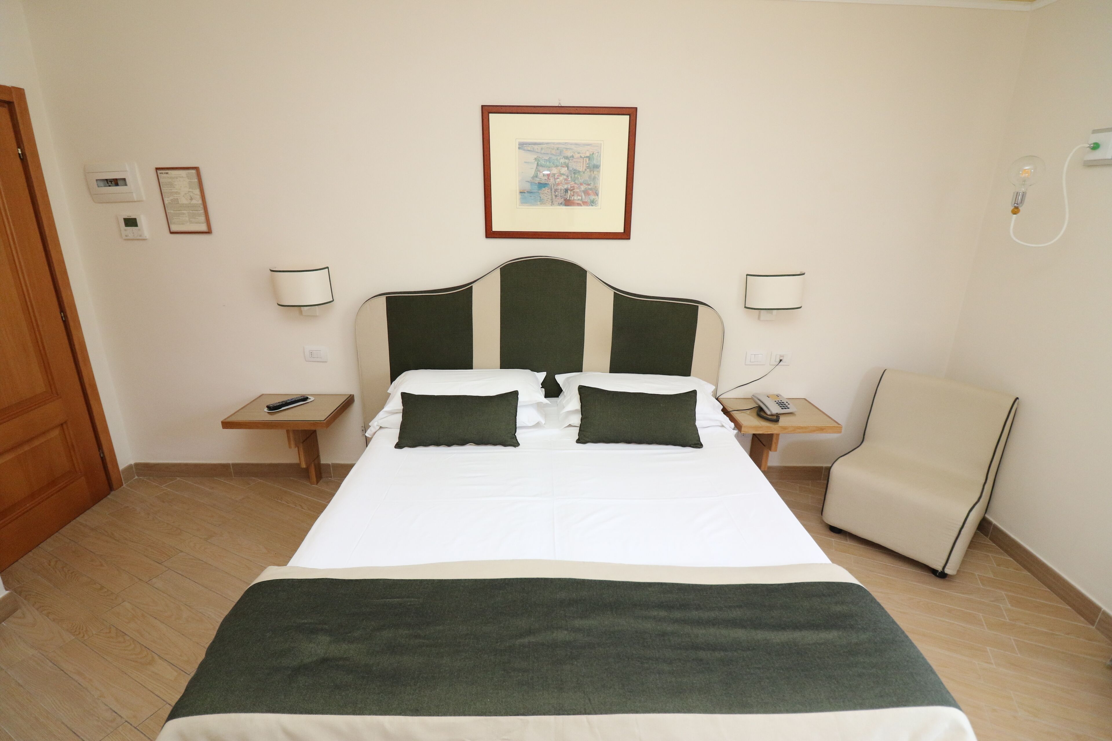 Une chambre d’hôtel avec un lit, deux tables de chevet, une chaise, un tableau encadré au mur et une prise électrique.